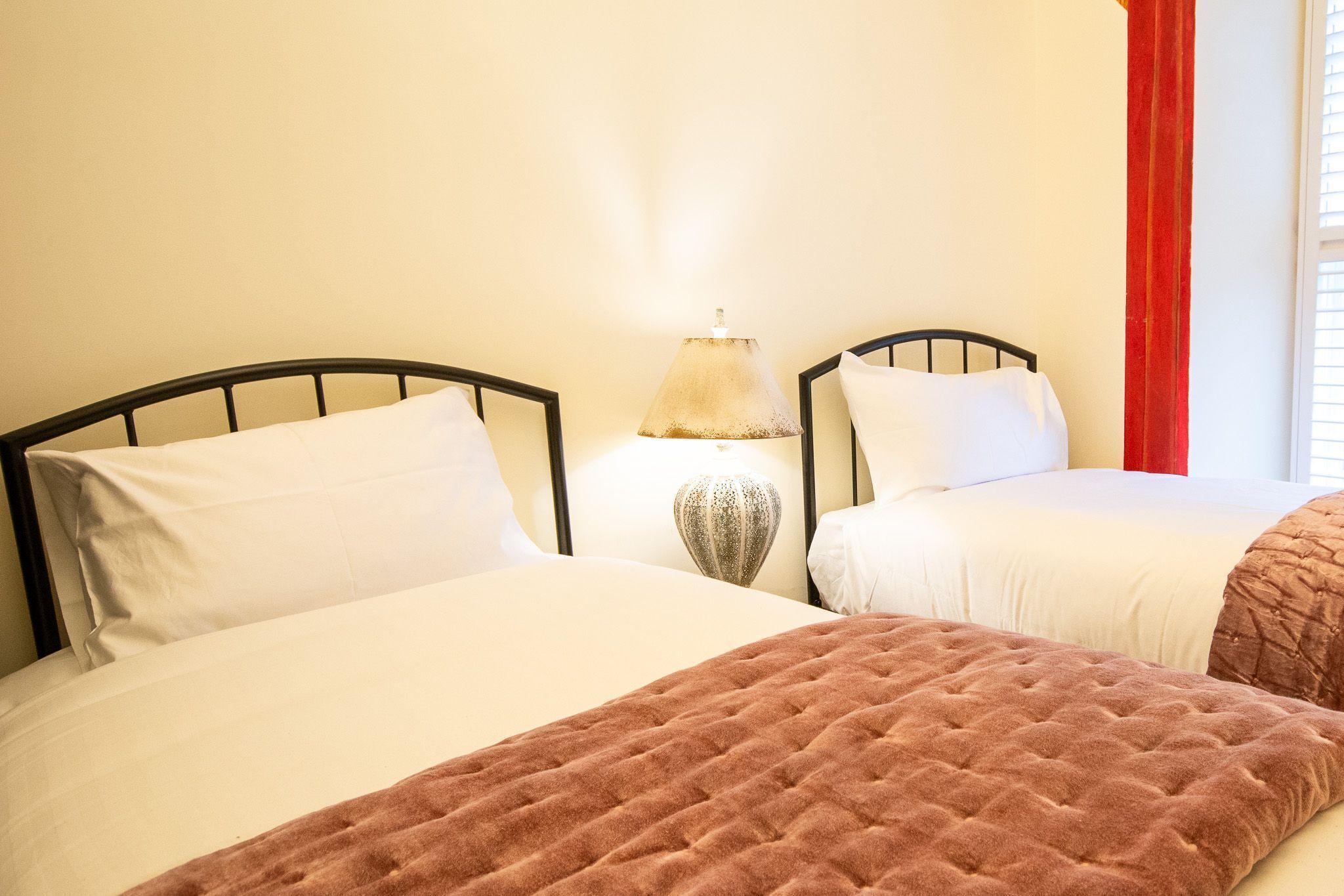 A hotel room with two beds, a lamp, and a window with red curtains.