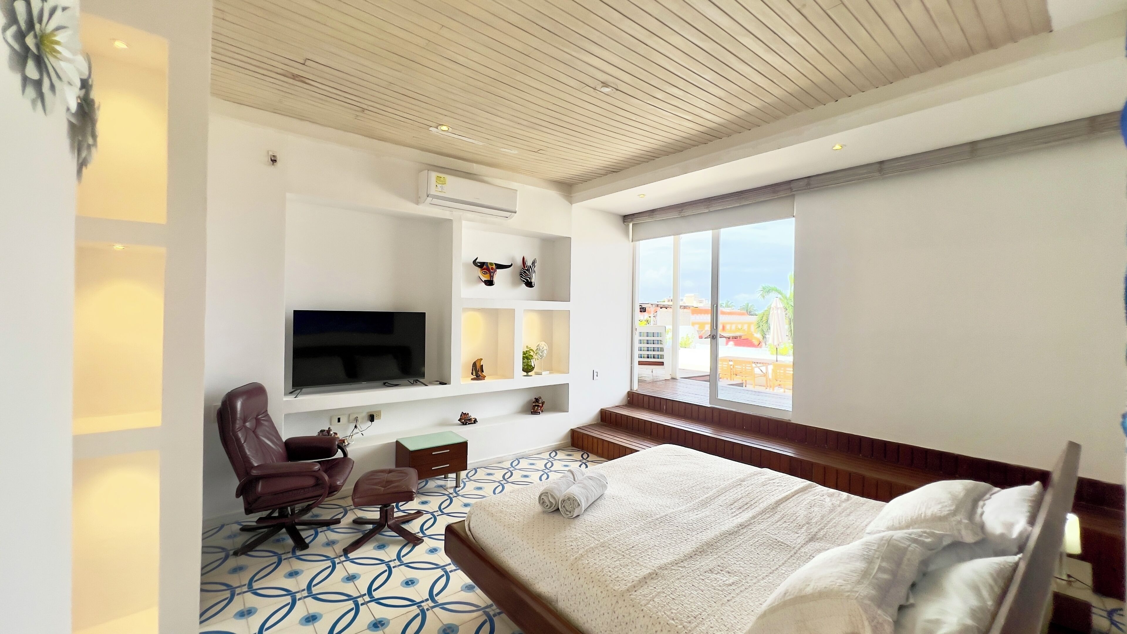 Habitación de hotel con cama, televisor montado en la pared, escritorio con silla y ventana con vista a edificios.