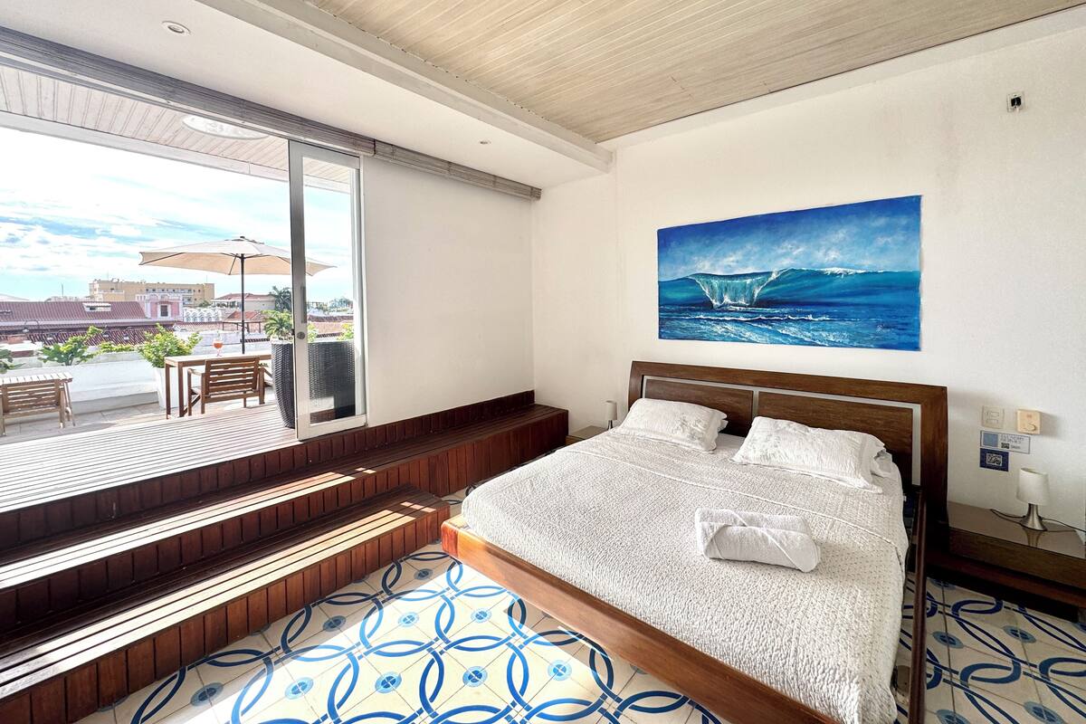 Une chambre d’hôtel avec un grand lit, une vue sur l’océan par une porte coulissante, un tableau représentant une vague et un sol carrelé.
