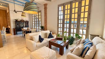 A spacious living room with a high ceiling, a dining area, and large windows with wooden frames.
