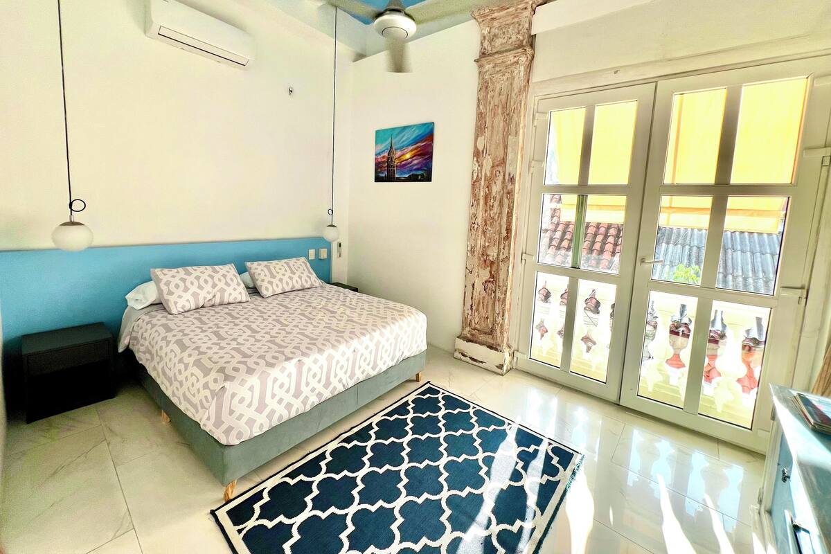 Une chambre à coucher avec un lit, un ventilateur de plafond, une fenêtre avec des rideaux et une vue sur l’extérieur.