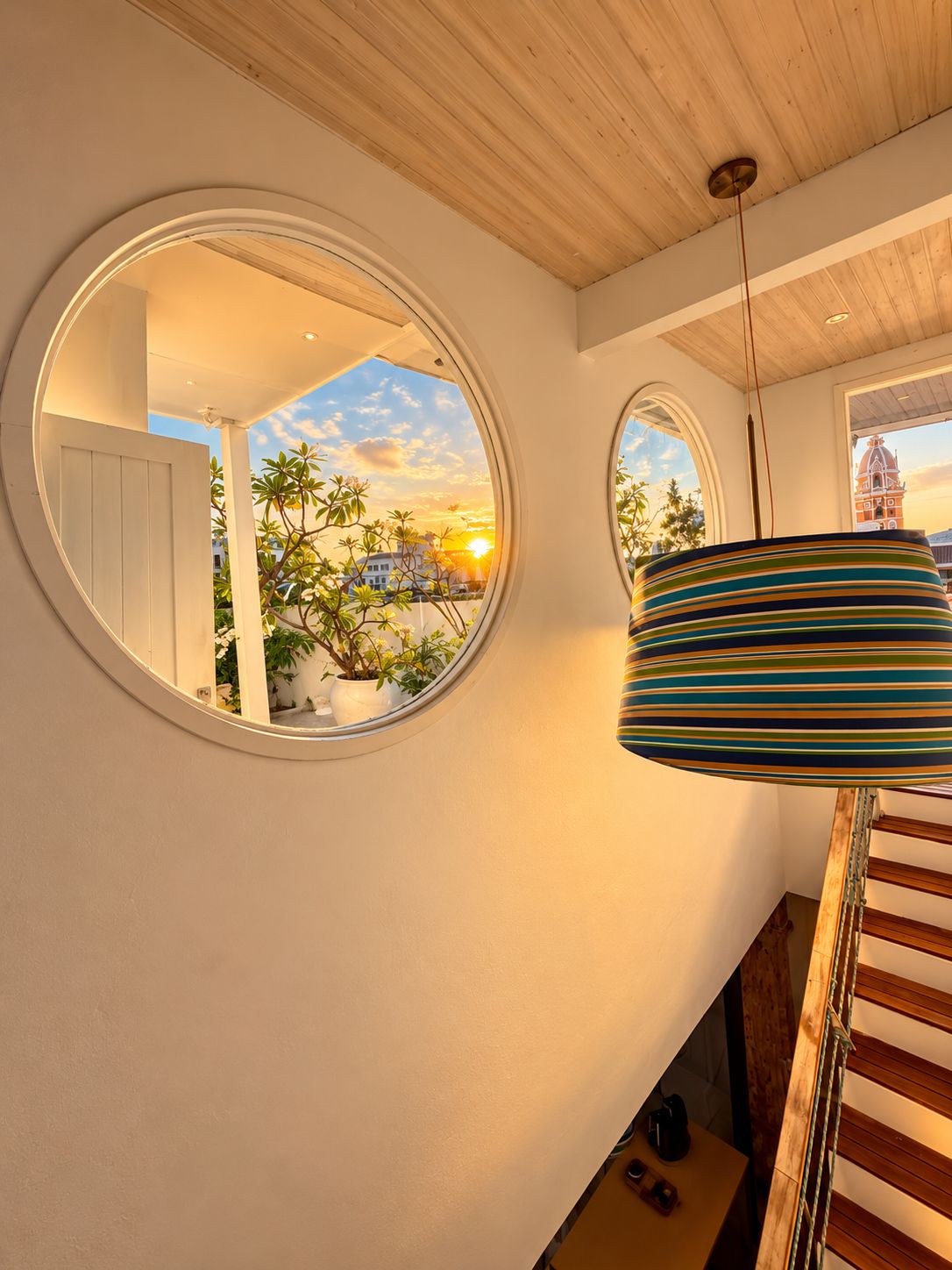 Un interior moderno con una escalera, un espejo redondo que refleja un atardecer y una lámpara a rayas.