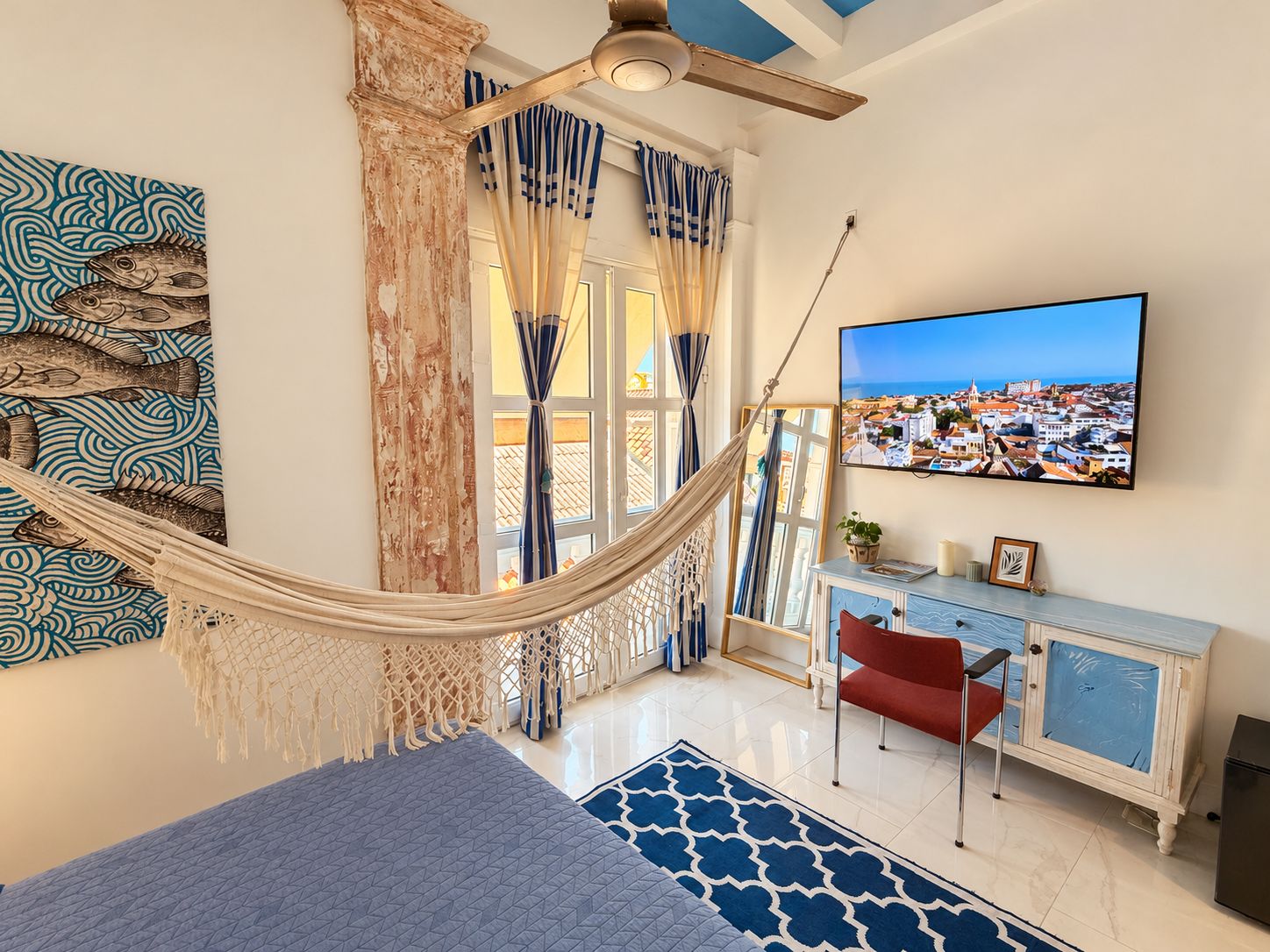 Una habitación con una hamaca, un ventanal con cortinas, un televisor y vistas a un pueblo costero.