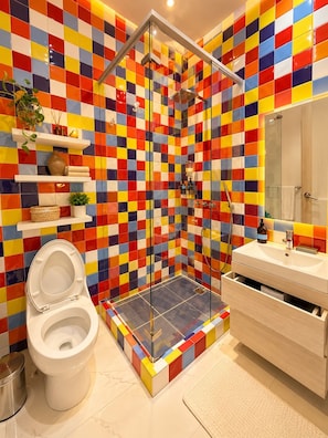 A bathroom with a colorful tiled wall, a white toilet, a glass-enclosed shower, and a white sink with a mirror. - Casa Lazuli 303 Cartagena (Cartagena)