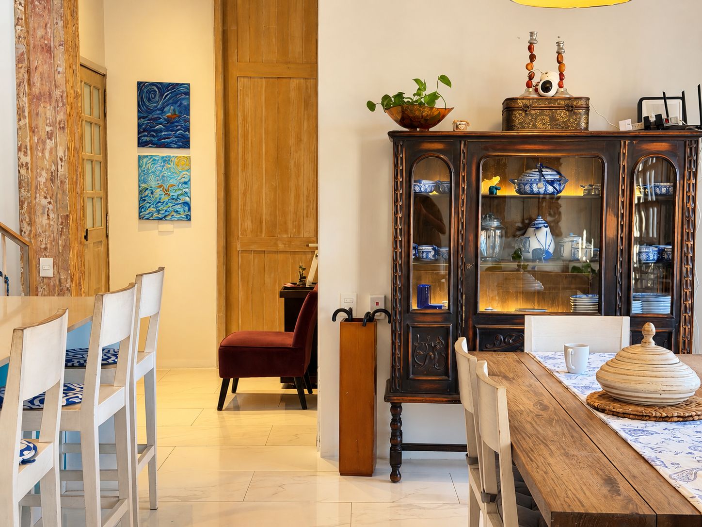 A dining area with a wooden table, white chairs, a red chair, a cabinet with blue and white dishes, and two paintings on the wall.