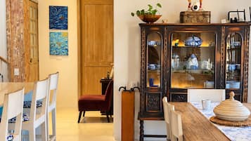 Un comedor con mesa de madera, sillas blancas, una silla roja, un armario con vajilla azul y blanca, y dos cuadros en la pared.