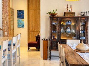 Een eetgedeelte met een houten tafel, witte stoelen, een rode stoel, een kast met blauw en wit servies, en twee schilderijen aan de muur.