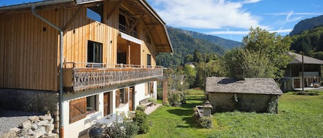A wooden chalet with a balcony, stone foundation, and a view of the surrounding greenery and mountains.