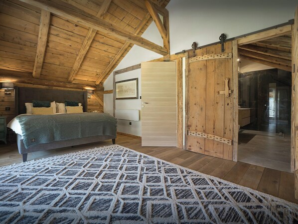 Une chambre à coucher avec un lit, un plafond en bois, une porte donnant sur une salle de bain et un tapis à motifs.