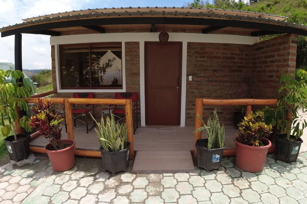 A small brick building with a wooden porch, potted plants, and a view of the surrounding landscape.