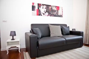 A modern living room with a black leather sofa, a patterned throw pillow, a small white side table with a lamp, and a large abstract painting on the wall.