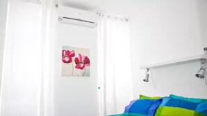 Un dormitorio con cama, cortinas blancas, un aire acondicionado de pared y un cuadro de flores.