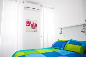A bedroom with a bed, white curtains, a wall-mounted air conditioning unit, and a painting of flowers.