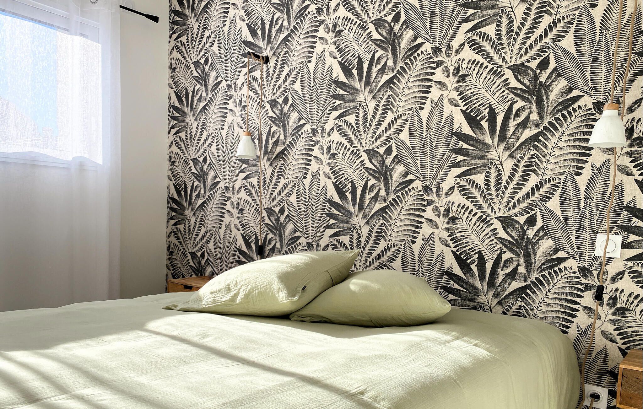 A bedroom with a bed, two pillows, a nightstand, a lamp, and a wall with a fern leaf pattern.