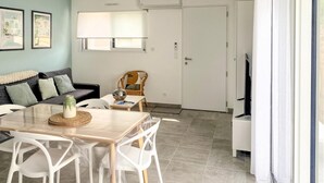 Ein modernes Wohnzimmer mit Essbereich, ausgestattet mit einer Couch, einem Couchtisch und einem Esstisch mit Stühlen. Eine Tür führt in einen anderen Raum, ein Fenster mit Jalousien ist ebenfalls vorhanden.