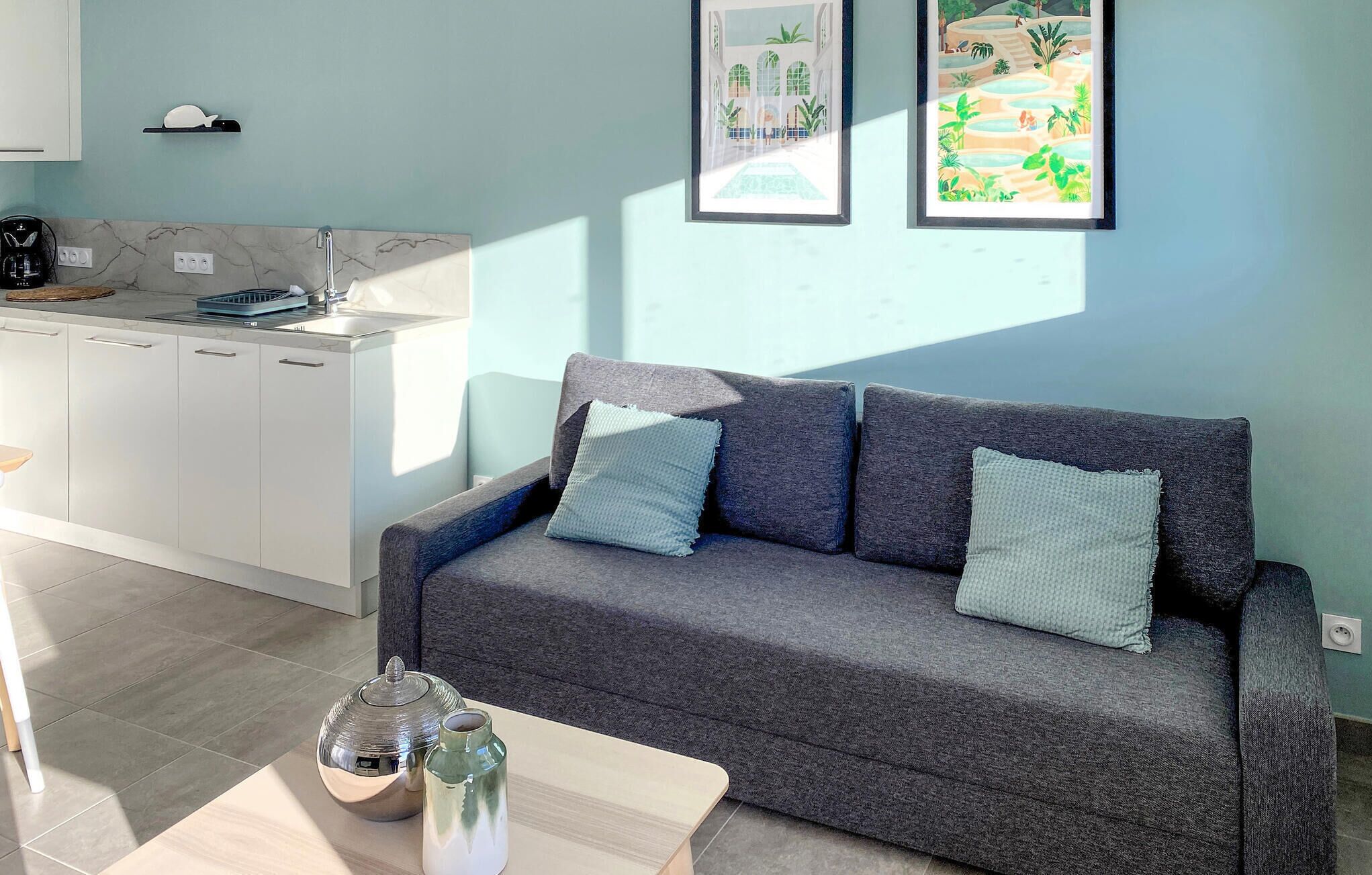 Een moderne woonkamer met een grijze bank, twee kussens, een bijzettafeltje, een waterkoker en een pot. Er is een keukenhoek met een wit aanrecht en een spoelbak. Twee ingelijste foto’s hangen aan de muur.