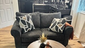 Una sala de estar moderna con un sofá gris, dos almohadones estampados, una mesa de centro redonda de madera con un jarrón y velas, y una cocina al fondo.