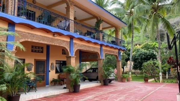 Un edificio de dos pisos con un balcón, plantas en macetas y un patio de tejas rojas.