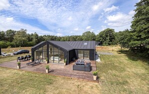 Ein modernes Haus mit einer großen Glasfassade, einer Terrasse mit Gartenmöbeln und einer umlaufenden Rasenfläche.