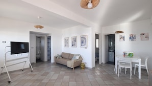 Un grand espace de vie comprenant un canapé, un téléviseur posé sur un meuble, une table à manger avec des chaises, et un coin cuisine visible à travers une porte ouverte.