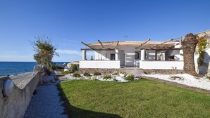 Une maison située sur la côte, avec un chemin de pierre menant à une porte blanche, une pelouse verdoyante et une vue sur la mer.
