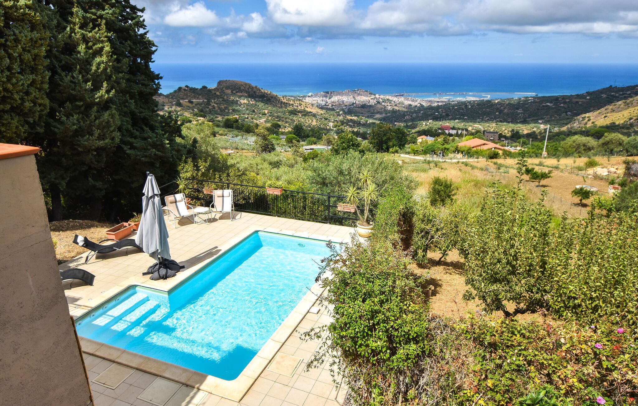 A swimming pool with lounge chairs and umbrellas, surrounded by greenery and a view of the sea and hills.
