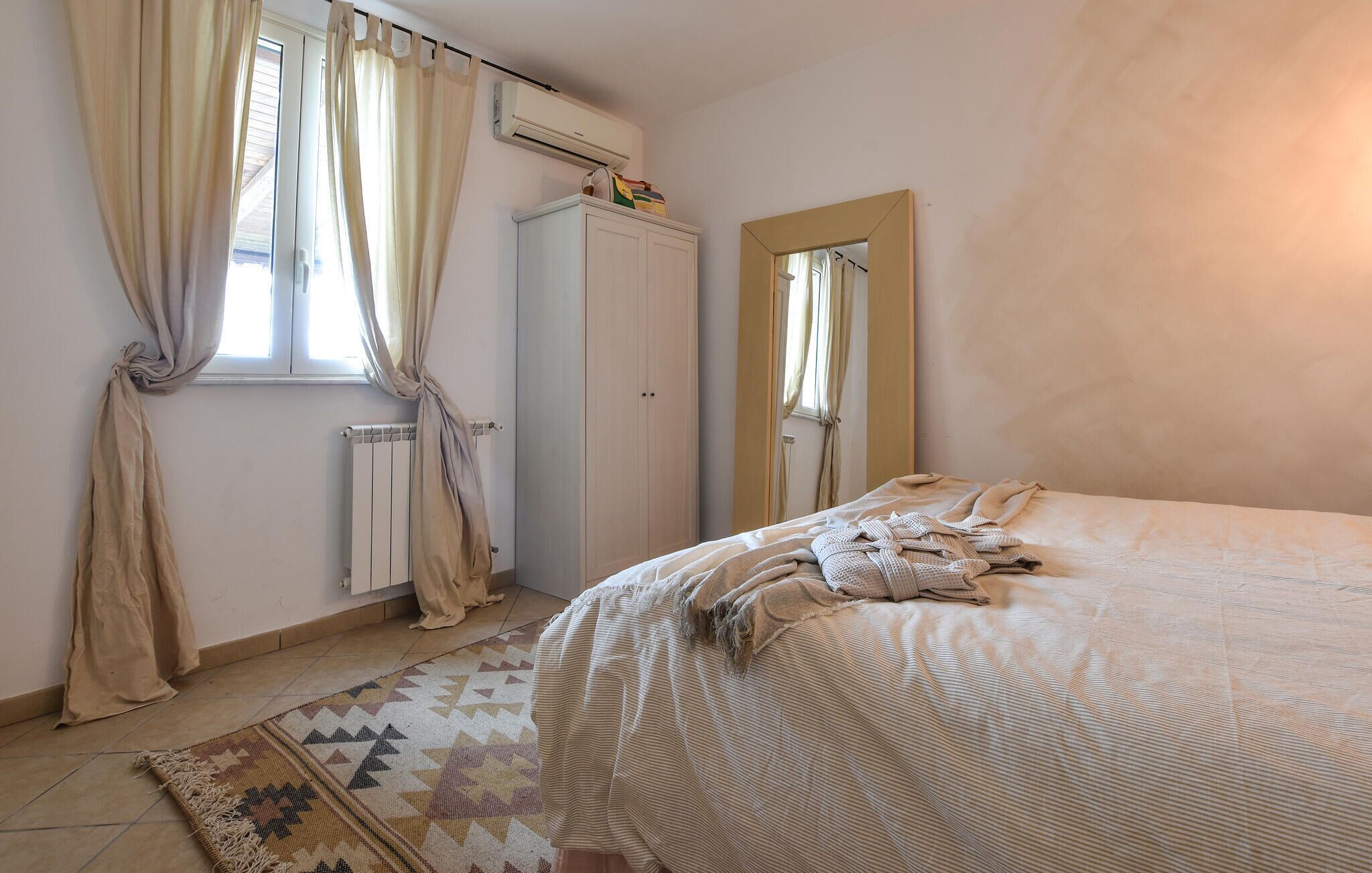 Un dormitorio con cama, radiador, armario, espejo y ventana con cortinas.