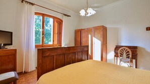 Un dormitorio con una cama, un armario de madera, un escritorio con una silla, un televisor y una ventana con cortinas.