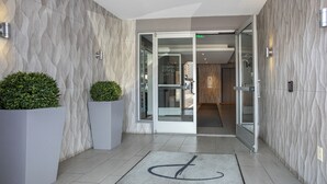 Ingresso moderno di un hotel con parete strutturata, porta in vetro e tappetino d'ingresso con simbolo.