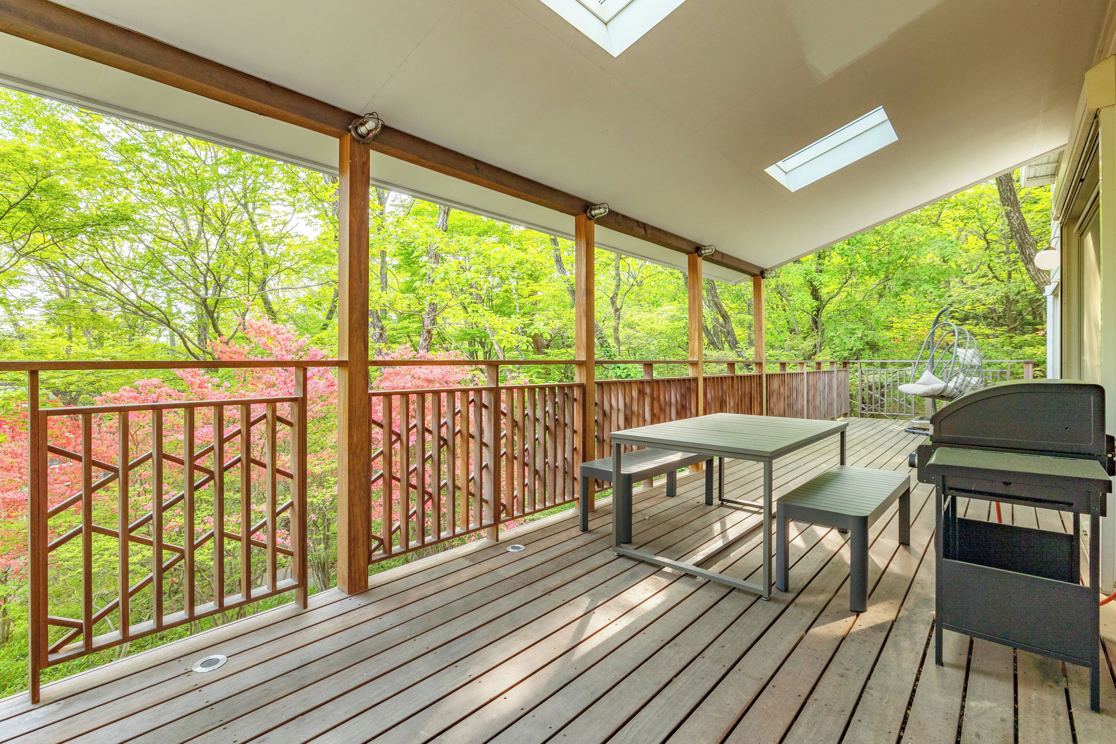 A covered outdoor deck with a table and chairs, a grill, and a view of trees and pink flowers.