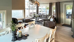 A modern living room with a dining table, white chairs, a sofa, and a large window overlooking a body of water.