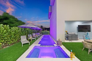 A modern outdoor pool area with a purple-lit water feature, surrounded by artificial grass and lounge chairs.