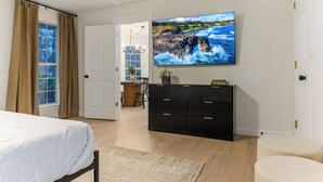 Ein Schlafzimmer mit einem Bett, einem an der Wand befestigten Fernseher, einer Kommode und einem Fenster mit Vorhängen.