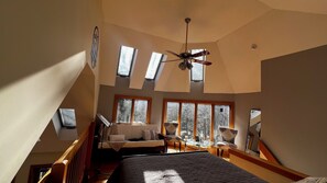 Une chambre avec un plafond haut, dotée de fenêtres de toit, comprenant un lit, un bureau et une chaise.