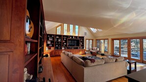 A spacious living room with a large sectional sofa, a wooden entertainment unit with a flat-screen TV, and large windows offering a view of the outdoors. - THE BIJOU -  a Precious Gem in the Green Mountains. HOT TUB, PET OK, HANDICAP (Rochester)