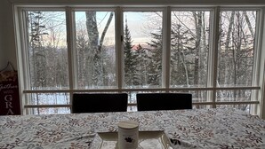 A room with a large window overlooking a forested area. There is a bed with a floral-patterned bedspread and a small table with a candle and a book. A sign on the wall reads "Gather with GRA." - THE BIJOU -  a Precious Gem in the Green Mountains. HOT TUB, PET OK, HANDICAP (Rochester)