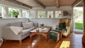 Un salon chaleureux avec un canapé, un fauteuil et un pouf, un plancher en bois et de grandes fenêtres ornées de plantes.