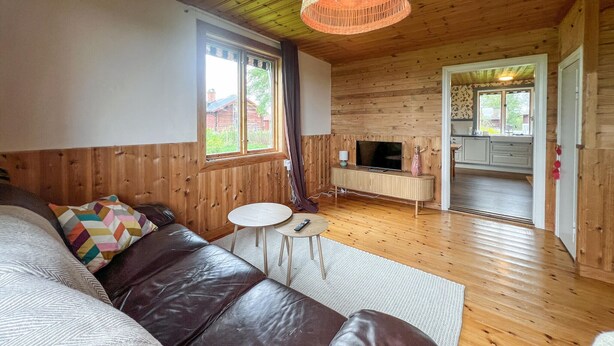 A cozy room with a leather sofa, a small round table, a TV, and a kitchen area visible through an open door. - Lovely home in Furudal with sauna (Furudal)