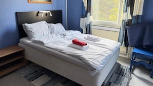 A neatly made bed with white linens and a red towel, a wooden nightstand, a framed picture on the wall, and a window with curtains. - Apartment in S West Sthlm close to Metro (Hägersten)