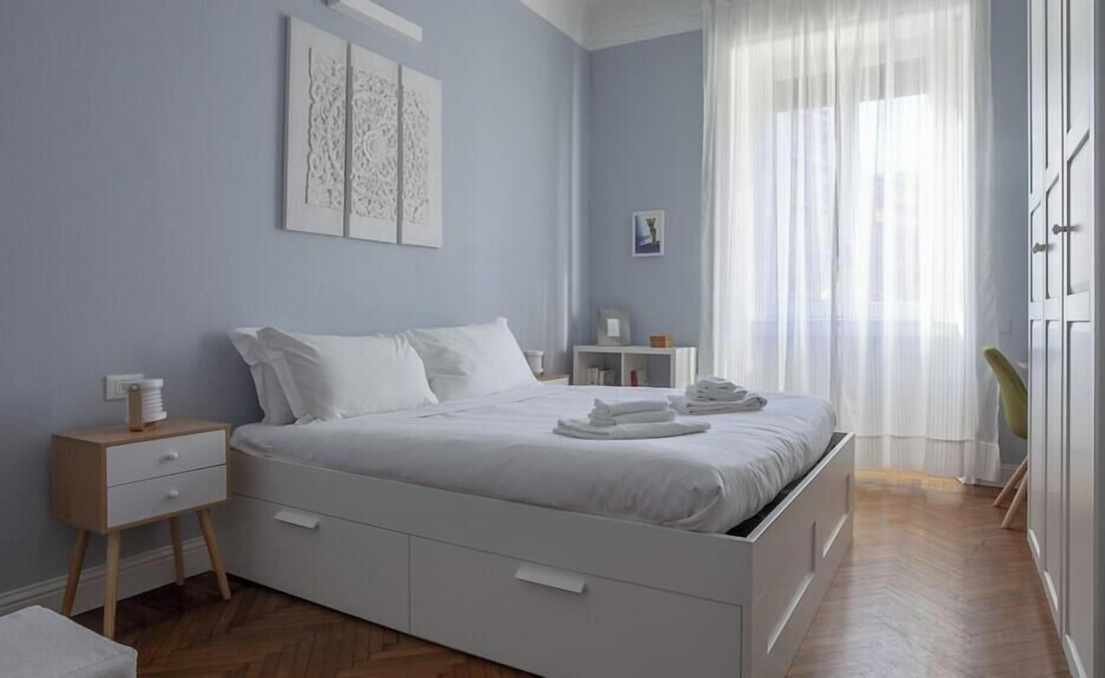 A bedroom with a bed, bedside tables, a nightstand, a chair, and a window with sheer curtains.
