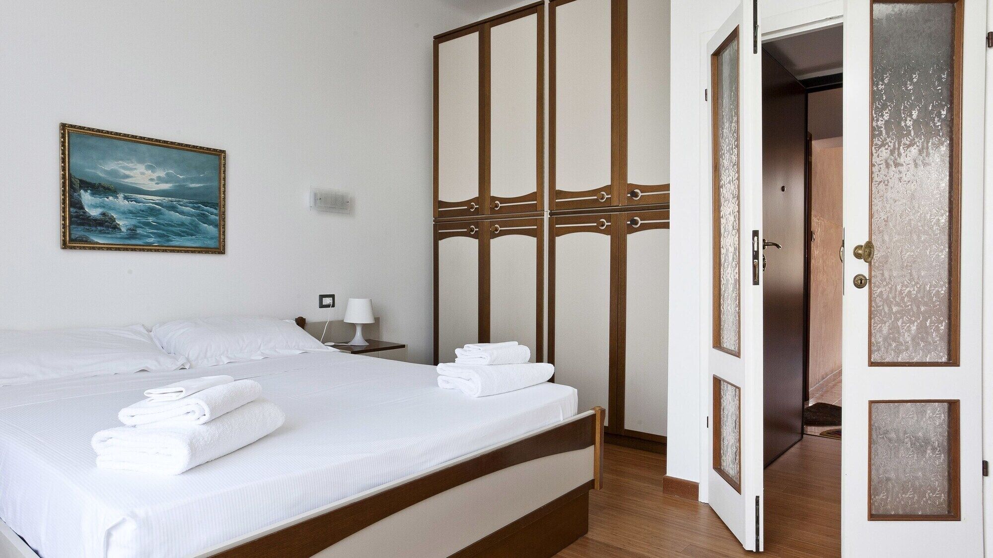 A hotel room with a bed, a nightstand, a lamp, a painting of the sea, and a wardrobe with a frosted glass door.