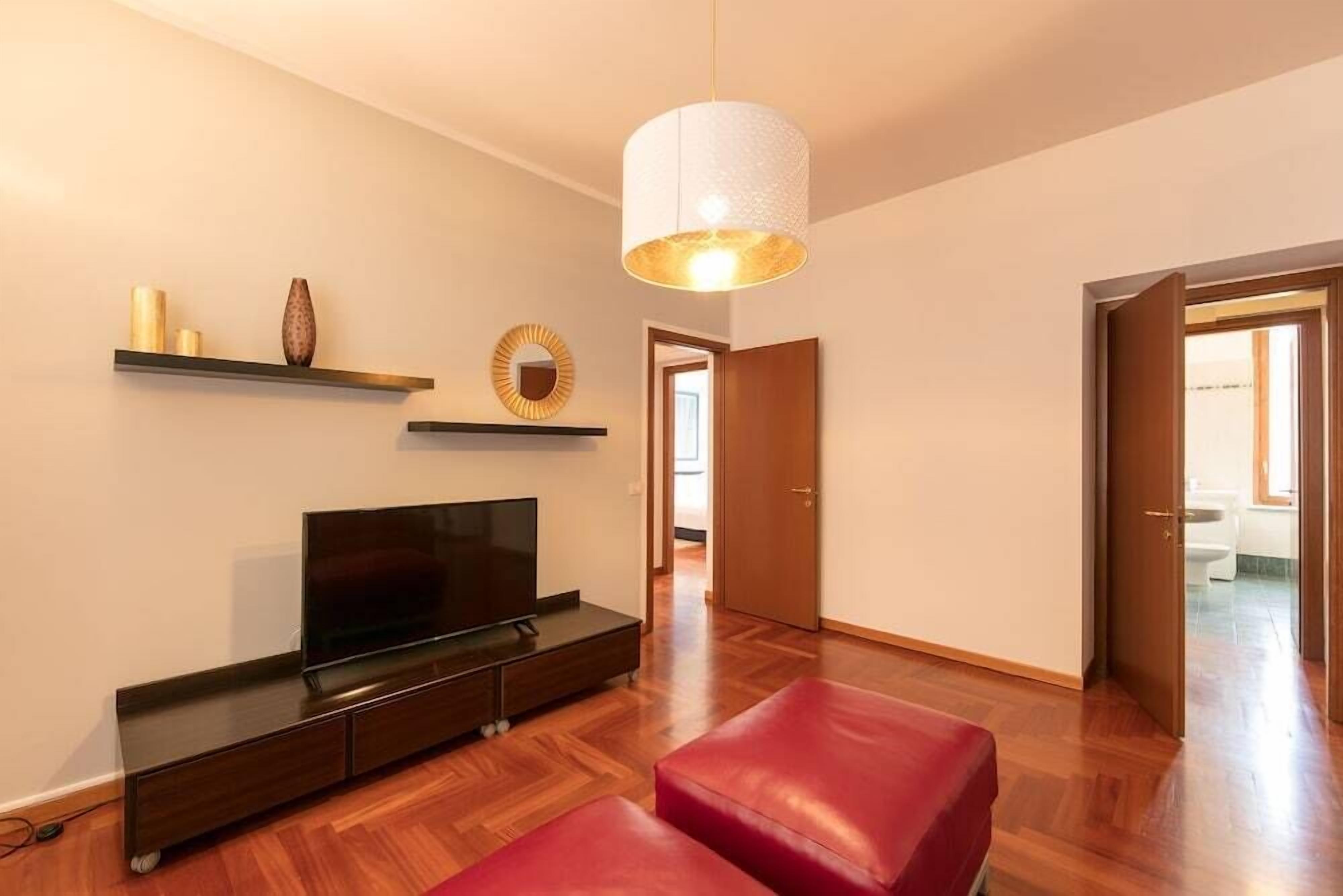 A modern living room with a flat-screen TV, wooden flooring, a red leather ottoman, and a pendant light.