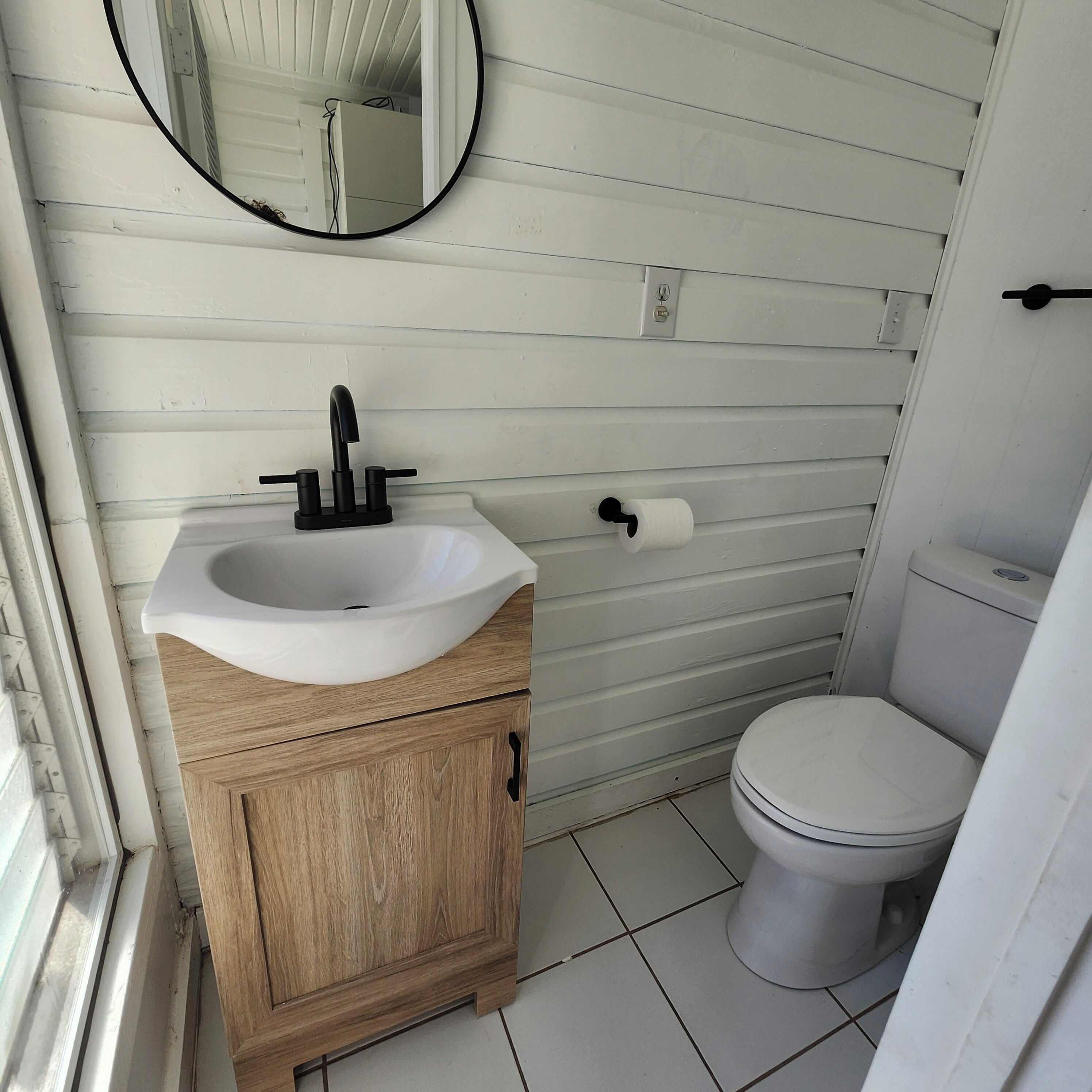 A bathroom with a white sink, wooden cabinet, toilet, and a round mirror on the wall.