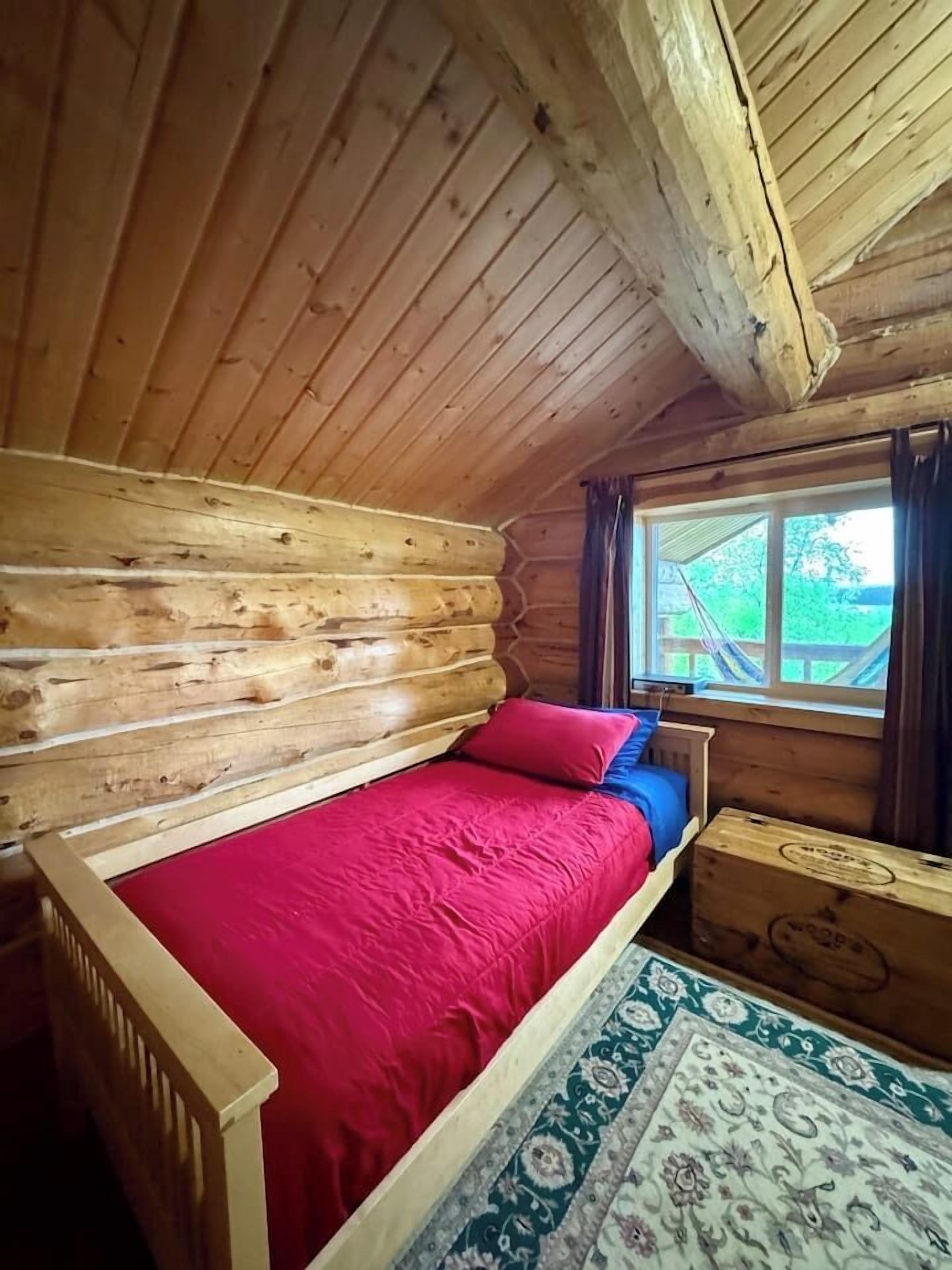 Una habitación de cabaña de madera con una cama, una ventana con cortinas y un pequeño cofre de madera.