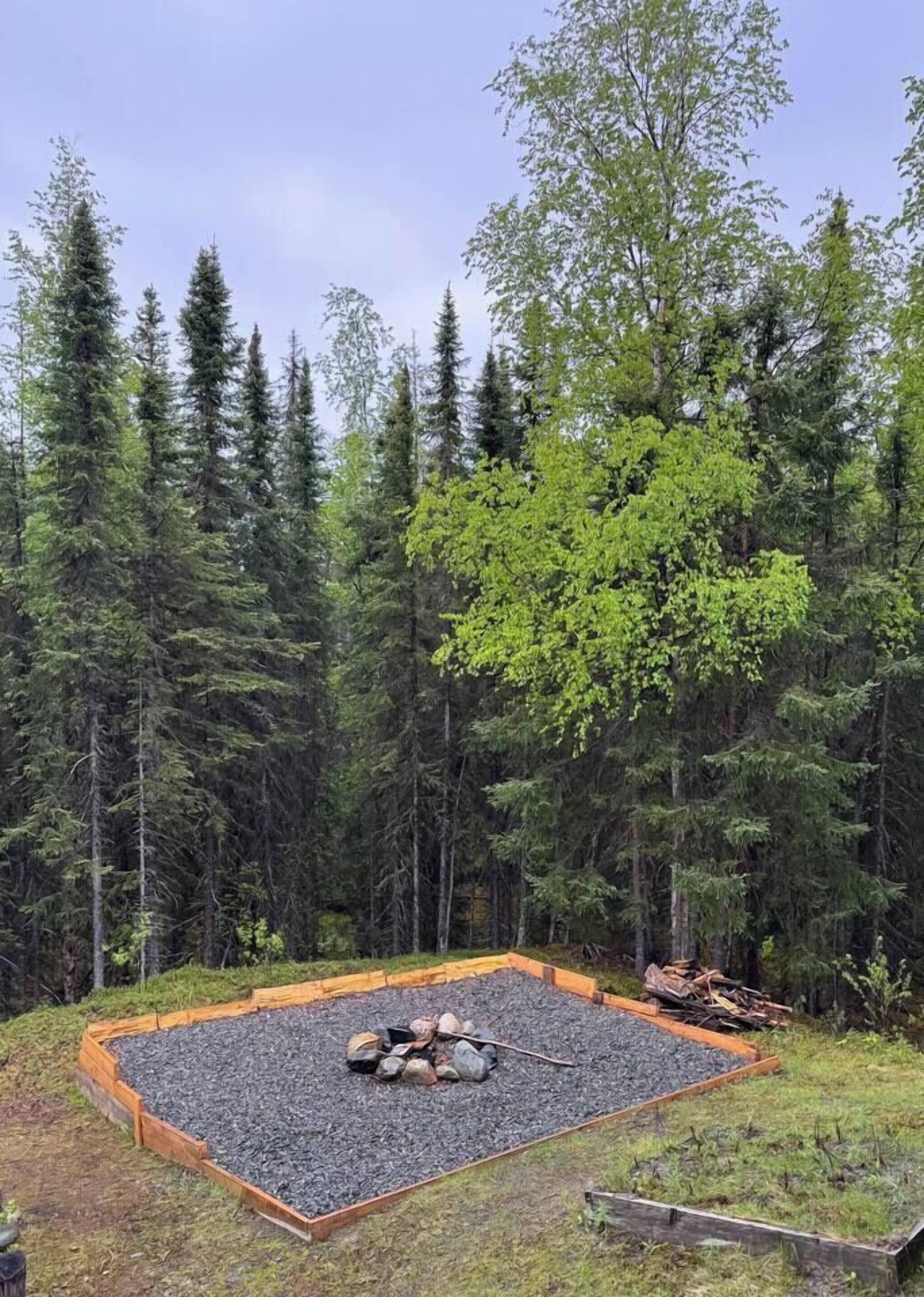 A square wooden deck with a fire pit in the center, surrounded by a forested area.
