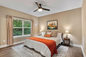 A bedroom with a bed, a ceiling fan, a window with curtains, a painting, two bedside tables with lamps, and a rug.