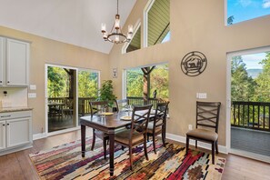 A dining area with a wooden table and chairs, a chandelier, and large windows offering a view of the outdoors.