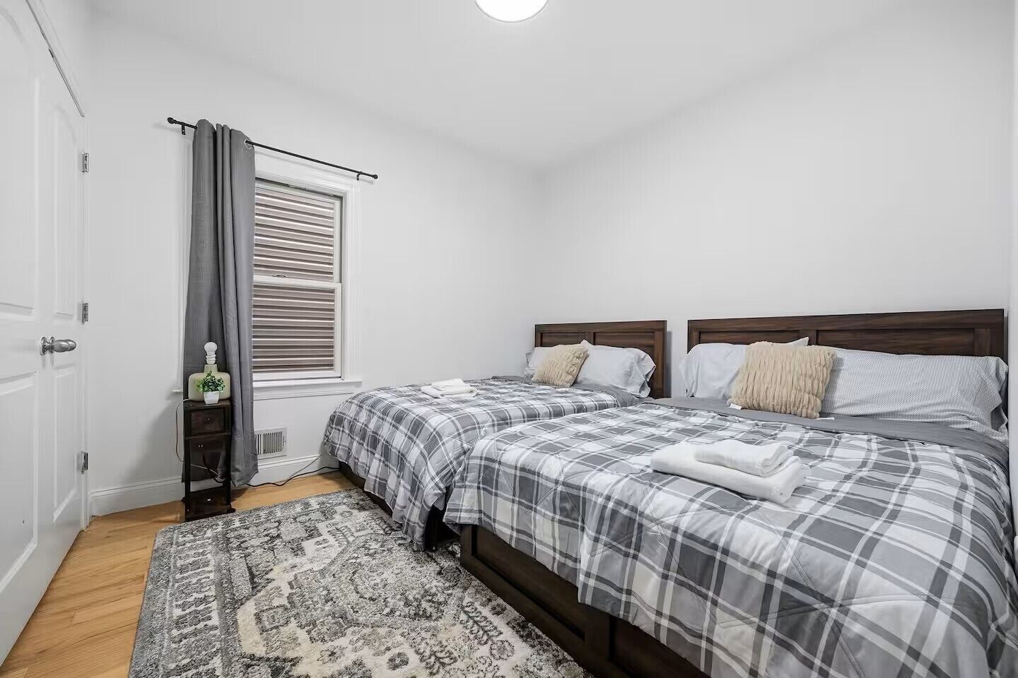 A bedroom with two beds, a window with blinds, a bedside table with a lamp, and a patterned rug.