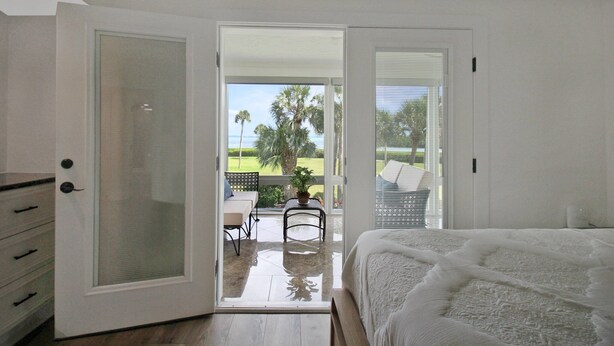 A bedroom with a large bed, a sliding glass door leading to a balcony with a view of palm trees and a lawn. - Luxe Waterfront Condo on Longboat Key - Beach Breeze (Longboat Key)