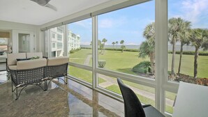 A room with a large window, a ceiling fan, a wicker sofa, and a view of a lawn and palm trees. - Luxe Waterfront Condo on Longboat Key - Beach Breeze (Longboat Key)